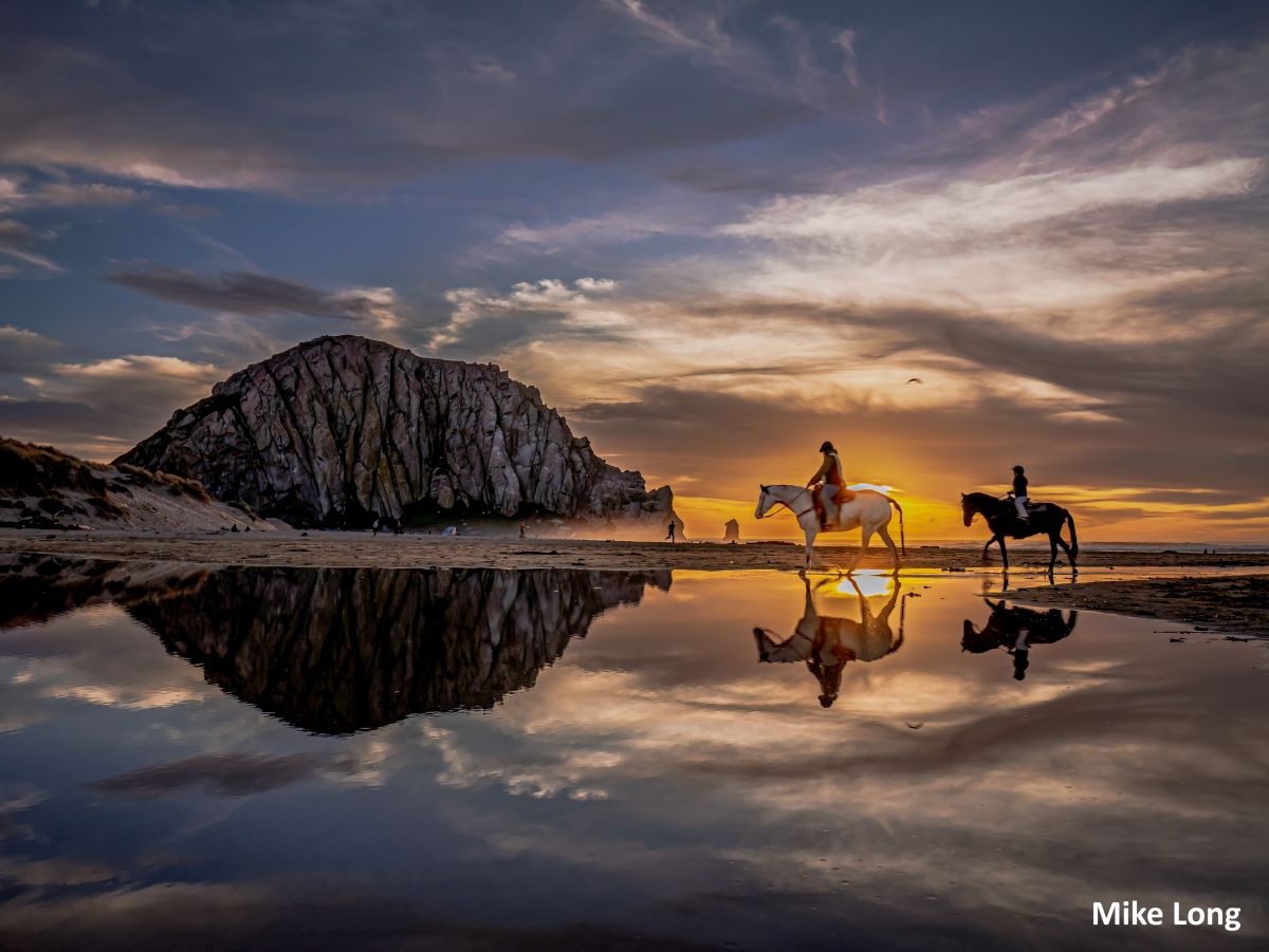Two people riding horses on the beach during sunset. Morro Rock reflects in the water alongside them.